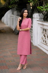 A woman modeling a pink textured kurta set with matching trousers, standing outdoors on a paved walkway next to a white decorative wall.