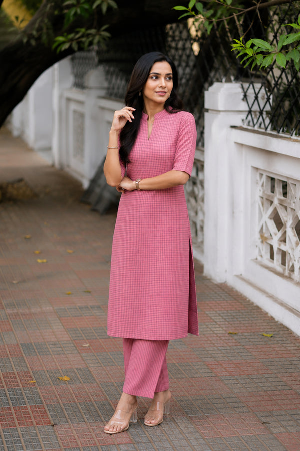 A woman modeling a pink textured kurta set with matching trousers, standing outdoors on a paved walkway next to a white decorative wall.